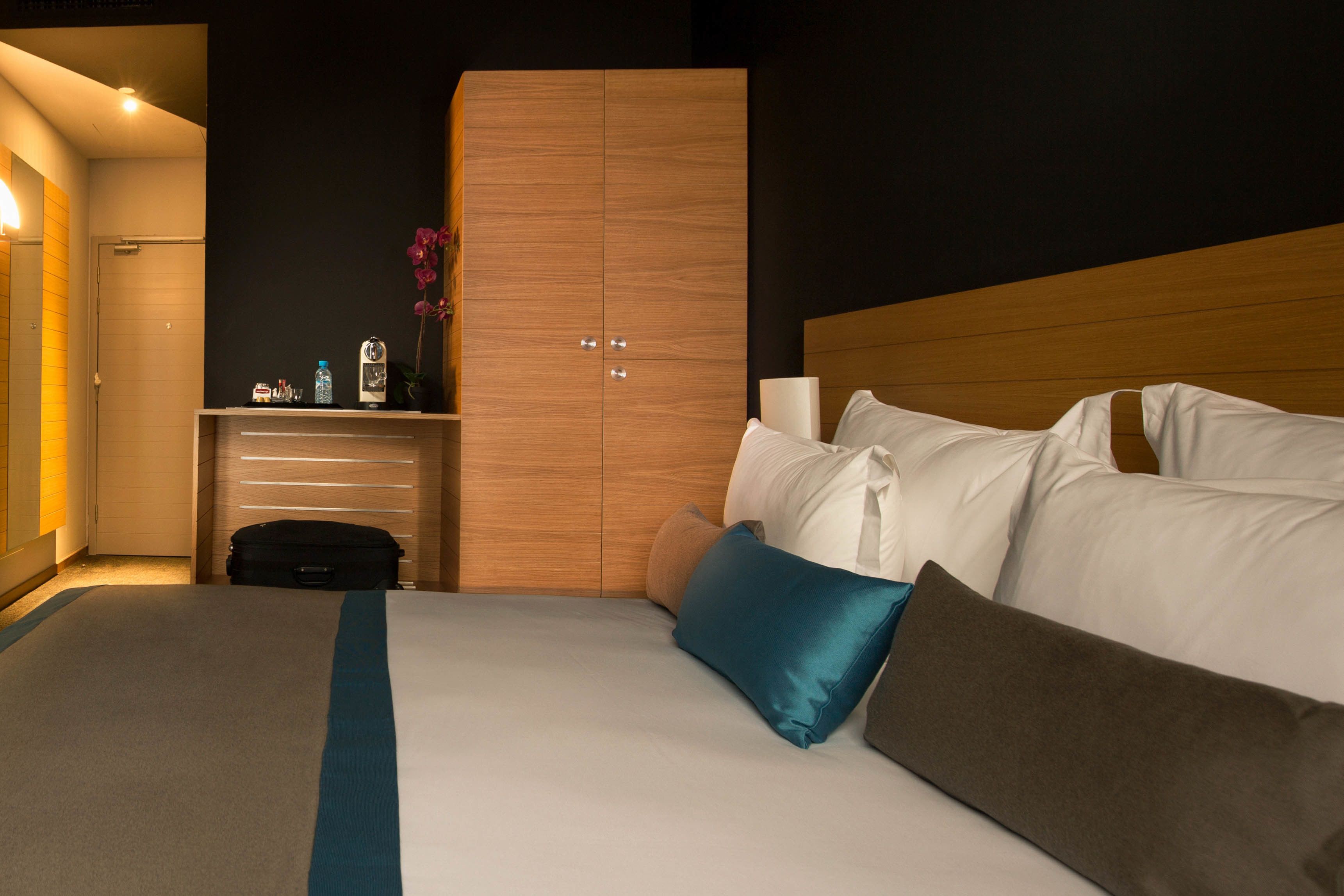 A hotel room with a bed, a wooden wardrobe, a bedside table with a lamp, and a suitcase.