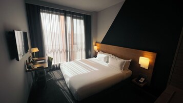 Una habitación de hotel con una cama grande, un escritorio con lámpara, una silla, una mesita de noche con lámpara y un ventanal con cortinas.