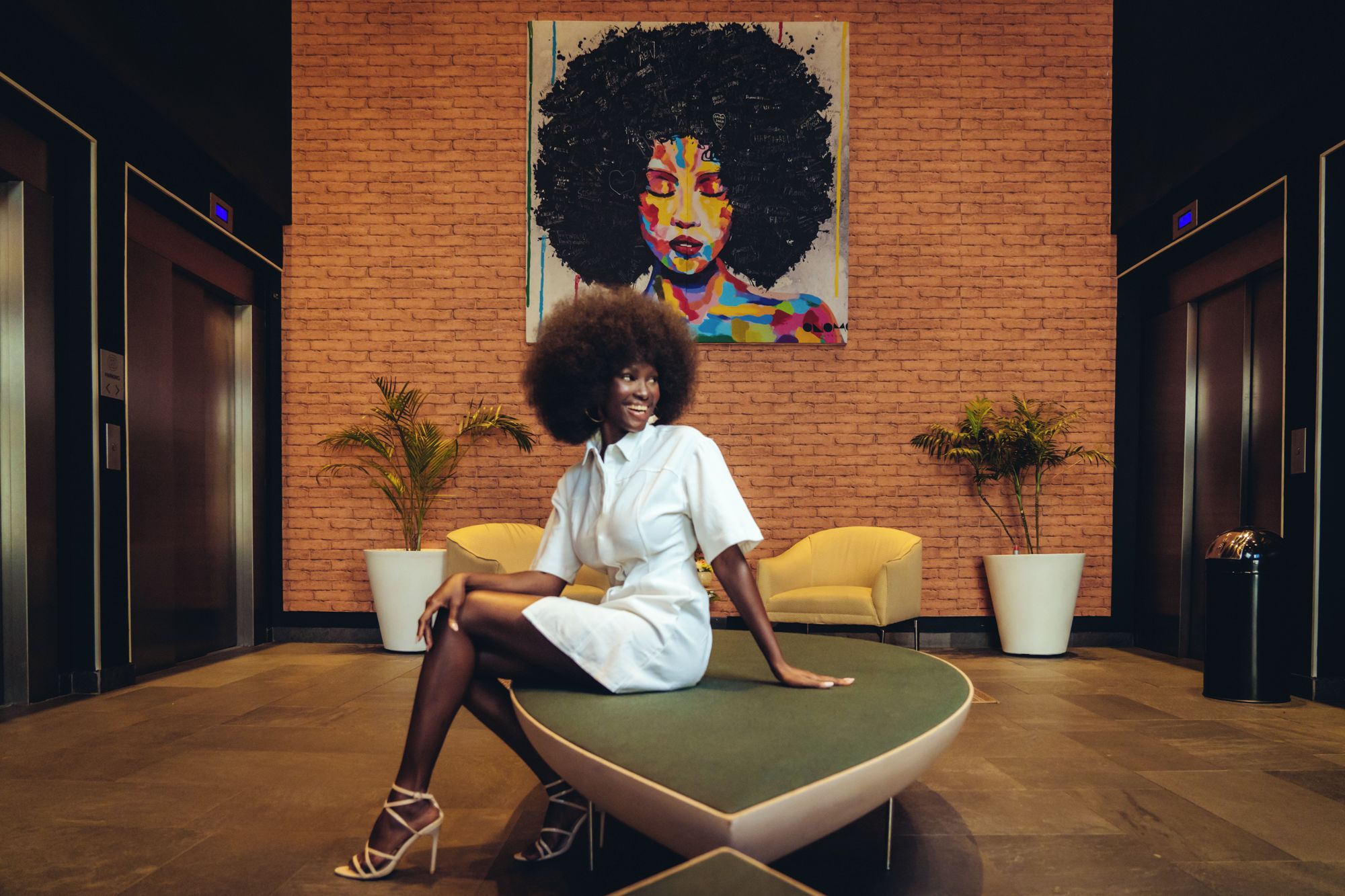 Une femme est assise sur une chaise verte dans un hall d’entrée, devant un mur de briques, avec deux plantes en pot et un tableau.