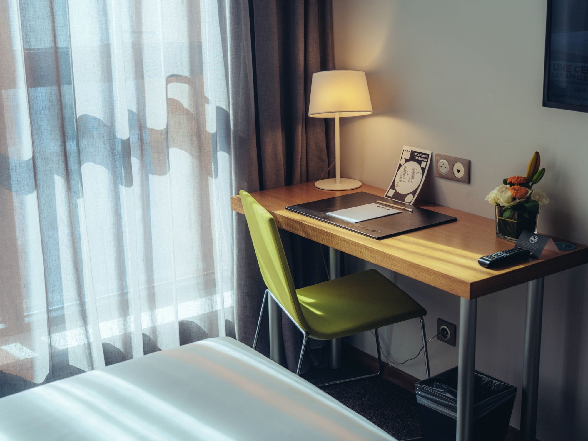 Hotellihuone, jossa on työpöytä, tuoli, lamppu, kaukosäädin ja kehyksiin asetettu televisio-ohjelmaopas.