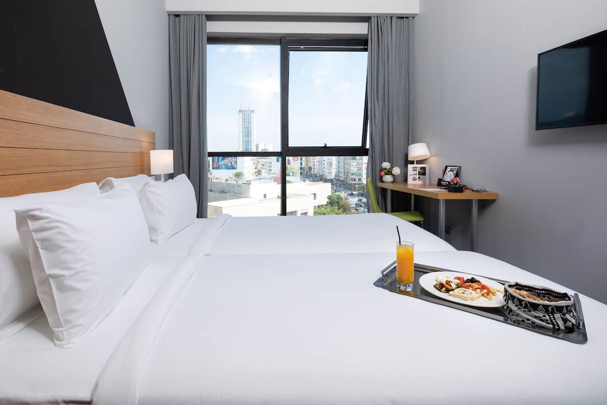 Une chambre d’hôtel avec un grand lit, une table de chevet avec une lampe, un plateau avec le petit-déjeuner, et une vue sur la ville par la fenêtre.