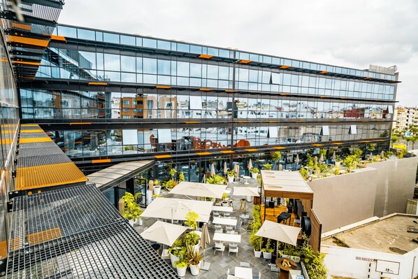 Un bâtiment moderne à plusieurs étages, doté d’espaces extérieurs aménagés avec des parasols et des plantes en pot.