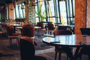 Un interior de restaurante con mesas y sillas redondas, ventanas amplias y plantas de interior.