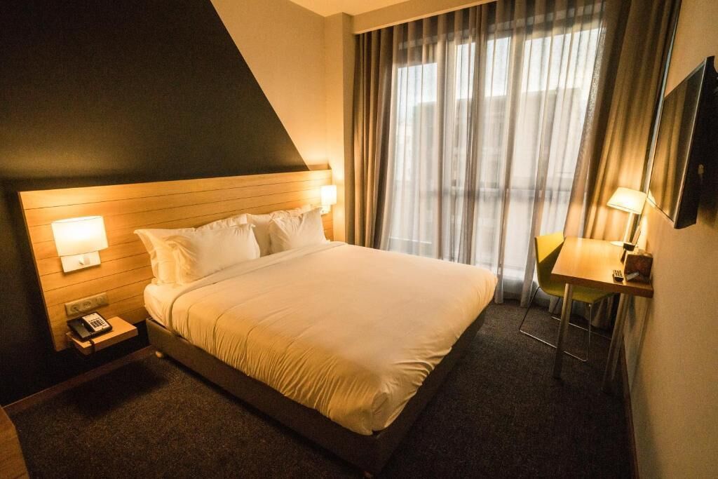 Quarto de hotel com cama, abajures, escrivaninha com cadeira e uma janela grande com cortinas.