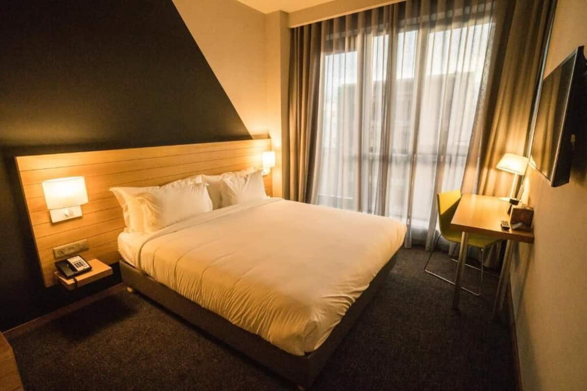 Une chambre d’hôtel avec un lit, des lampes de chevet, un bureau avec une chaise et une grande fenêtre avec des rideaux.