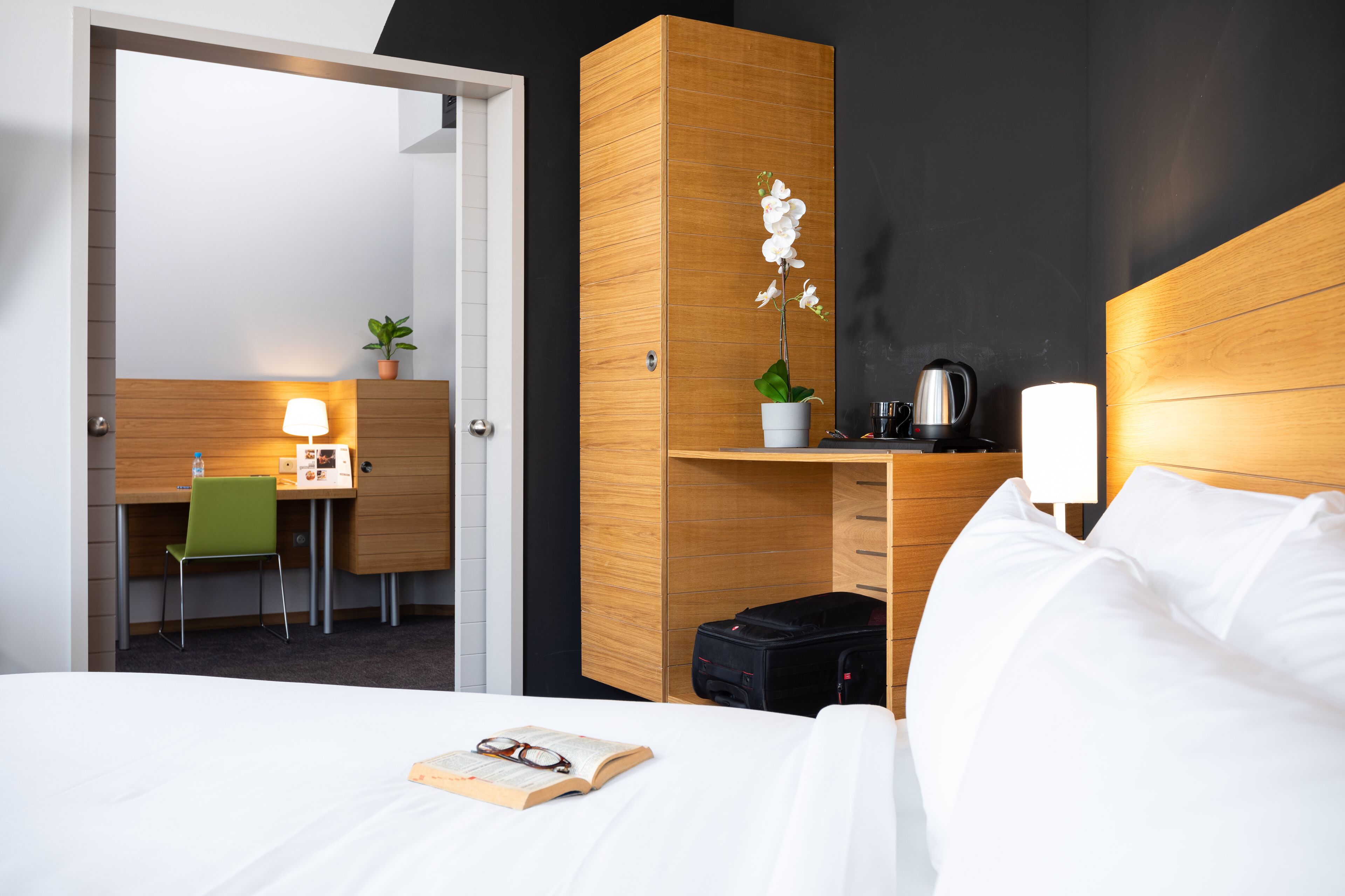 Une chambre d’hôtel avec un lit, un bureau et une chaise, une petite plante, une bouilloire et une valise.