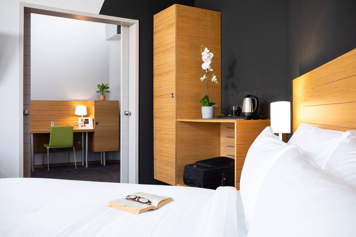 Une chambre d’hôtel avec un lit, un bureau et une chaise, une petite plante, une bouilloire et une valise.