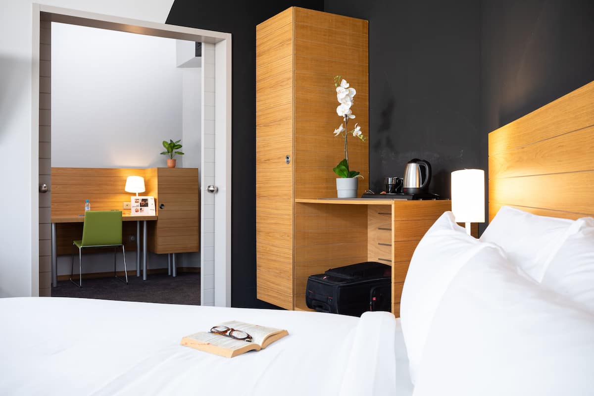 Une chambre d’hôtel avec un lit, un bureau et une chaise, une petite plante, une bouilloire et une valise.