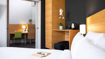Ett hotellrum med en säng, ett skrivbord med en stol, en liten växt, en vattenkokare och en resväska.