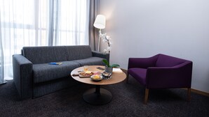 A modern hotel room with a grey sofa, a purple armchair, a round table with food and a lamp, and a potted orchid.