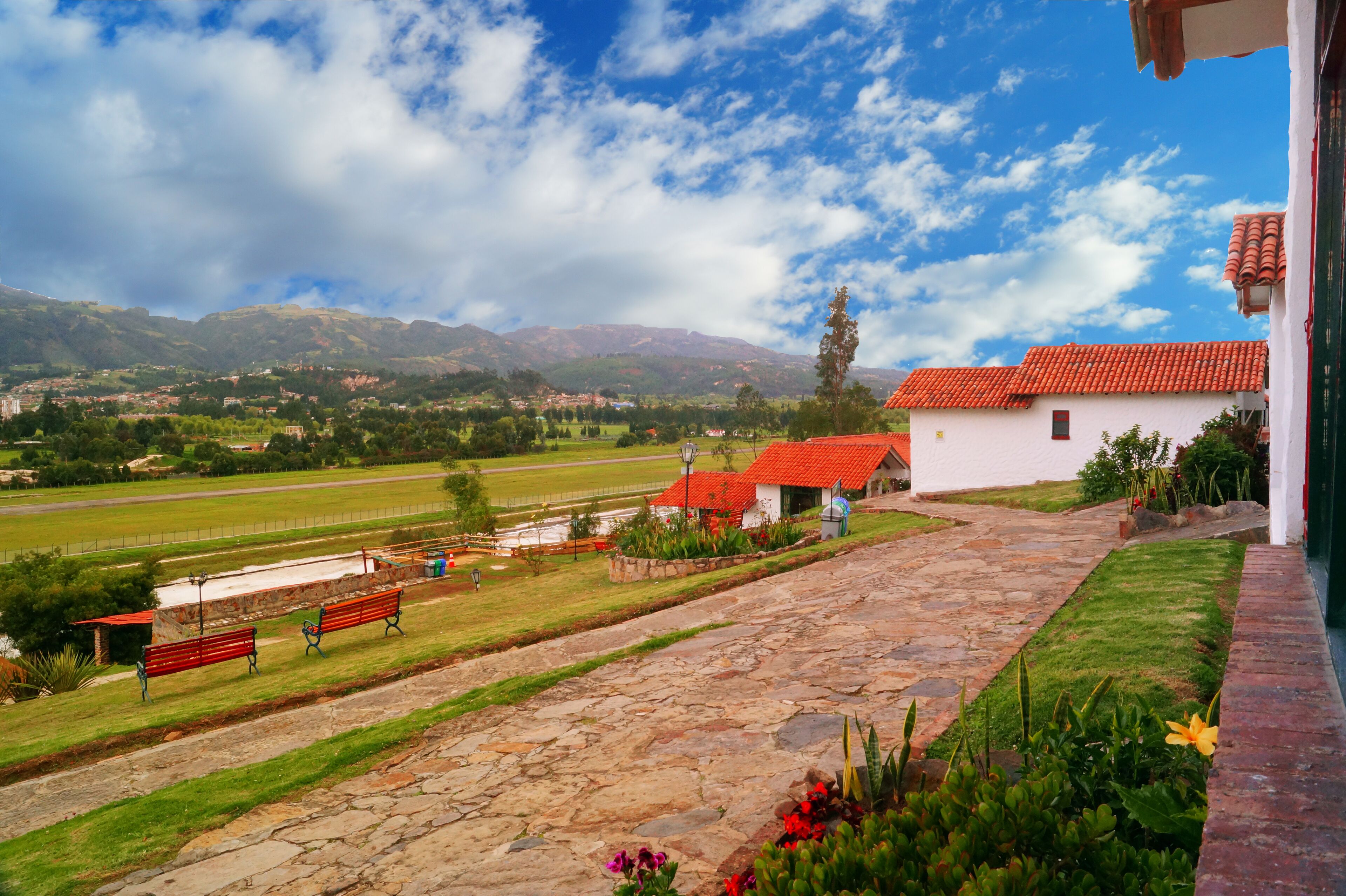 A stone pathway leading to a white building with a red roof, surrounded by a well-maintained garden and a view of a green field and mountains in the distance.