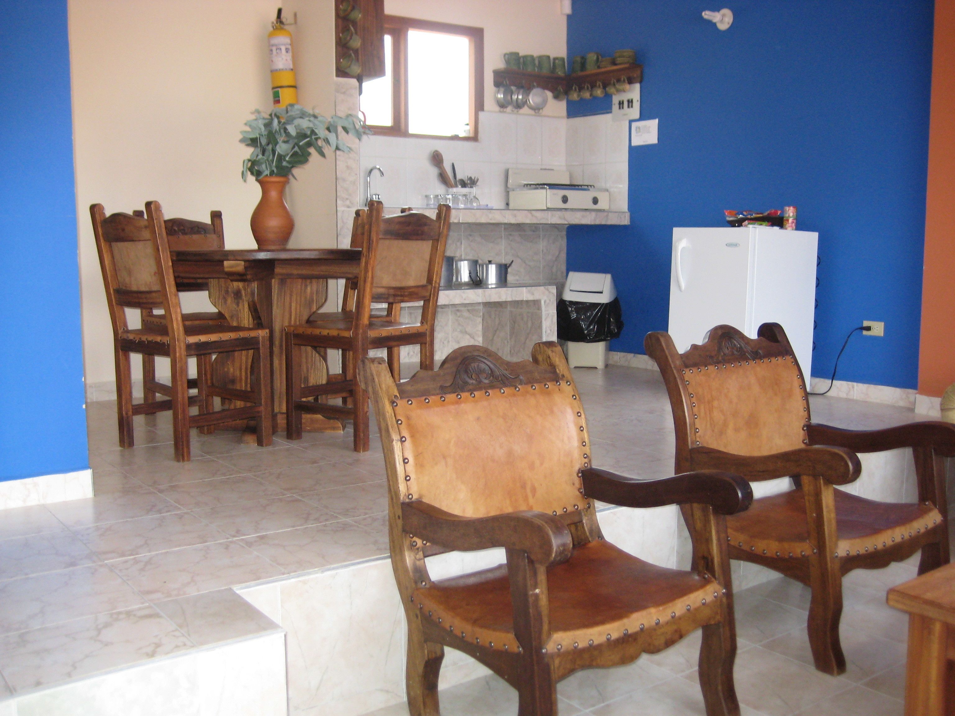 Un coin repas avec des chaises et une table en bois, une cuisine équipée d’un évier et d’une cuisinière, et un mur bleu.