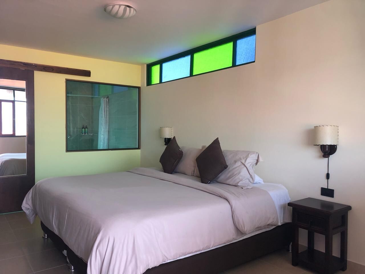 A bedroom with a large bed, two bedside tables, a nightstand, a glass shower enclosure, and a wall-mounted lamp.