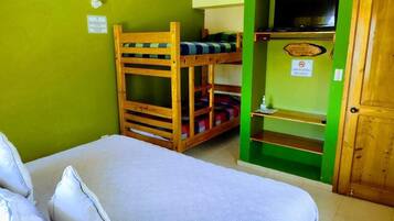 A room with a bunk bed, a single bed, a green wall, a wooden door, and a shelf with a television.