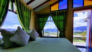 A bed with a quilted mattress and two pillows, a window with green curtains, and a view of a landscape through the window. - Hotel Cabañas San Cayetano (Paipa)