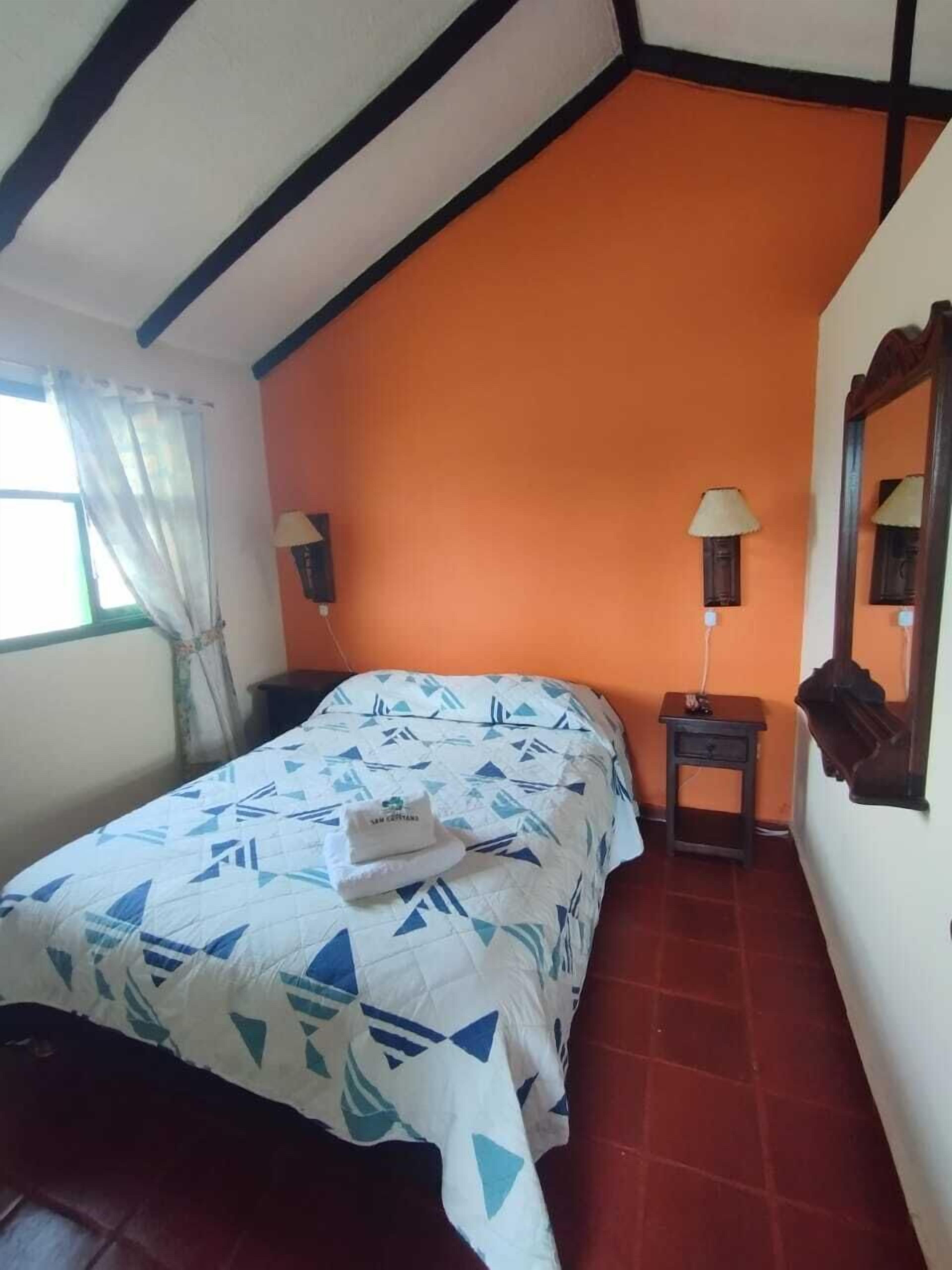 Un dormitorio con techo inclinado, una cama con colcha estampada, dos lámparas de noche y una mesita con lámpara.