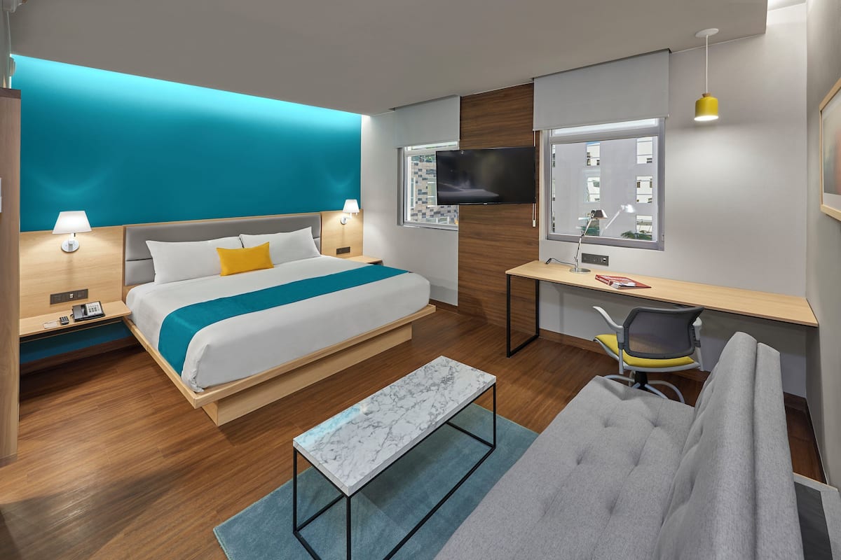 Ein modernes Hotelzimmer mit einem großen Bett, einem Schreibtisch, einem Sessel, einem Fernseher und einem Badezimmerspiegel.