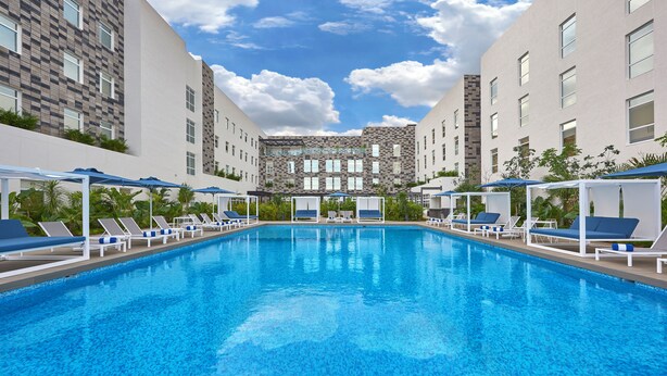 A swimming pool with lounge chairs and umbrellas, surrounded by buildings with a checkered facade and a stone wall in the background. - City Express Plus by Marriott Cancún Aeropuerto Riviera (Cancun)