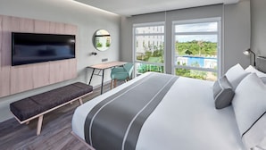 A modern hotel room with a large bed, a bench, a desk, a TV mounted on the wall, and large windows offering a view of a pool and greenery. - City Express Plus by Marriott Cancún Aeropuerto Riviera (Cancun)