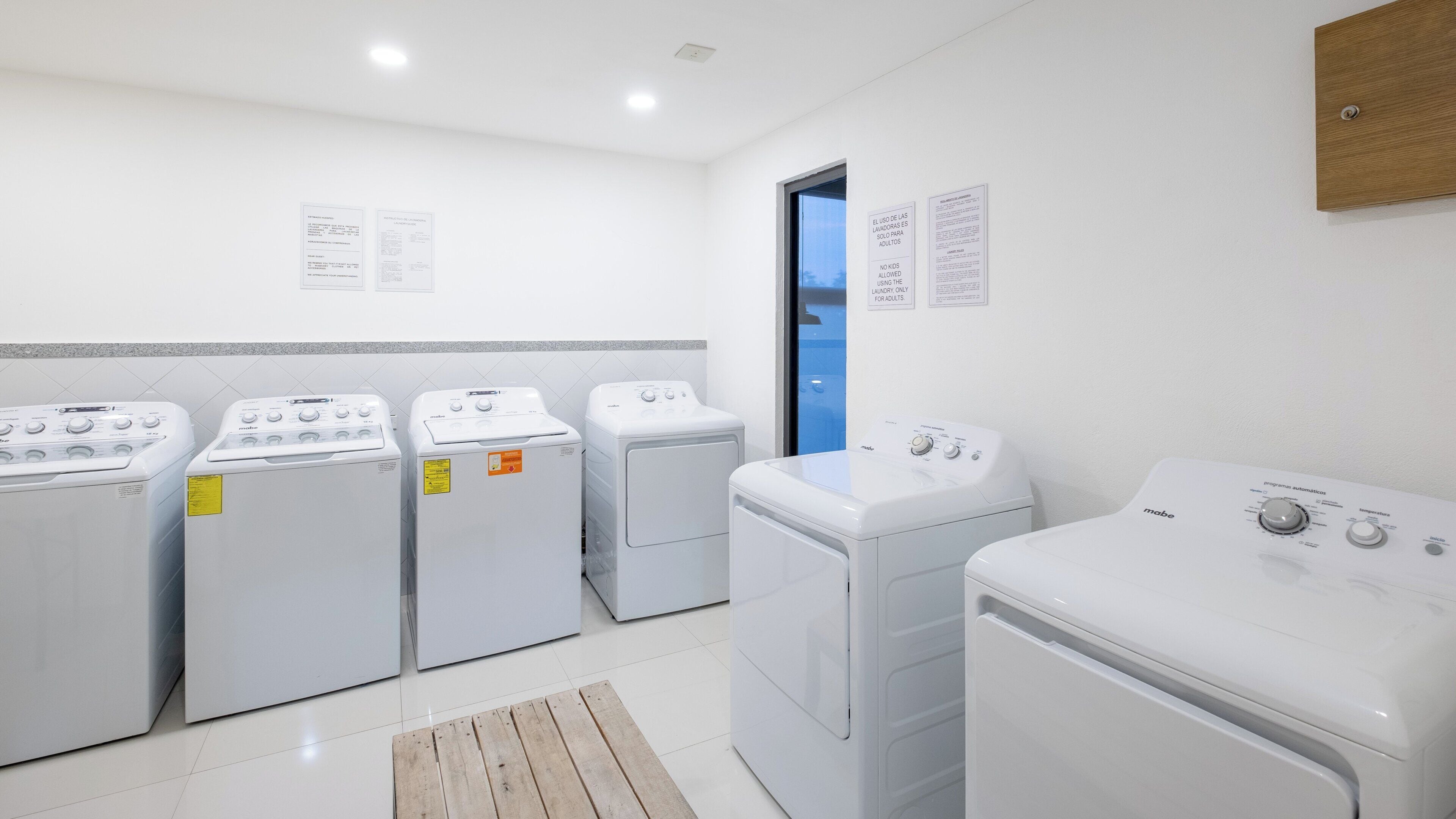 A laundry room with four front-loading washers and dryers, a wooden bench, and informational signs on the wall.