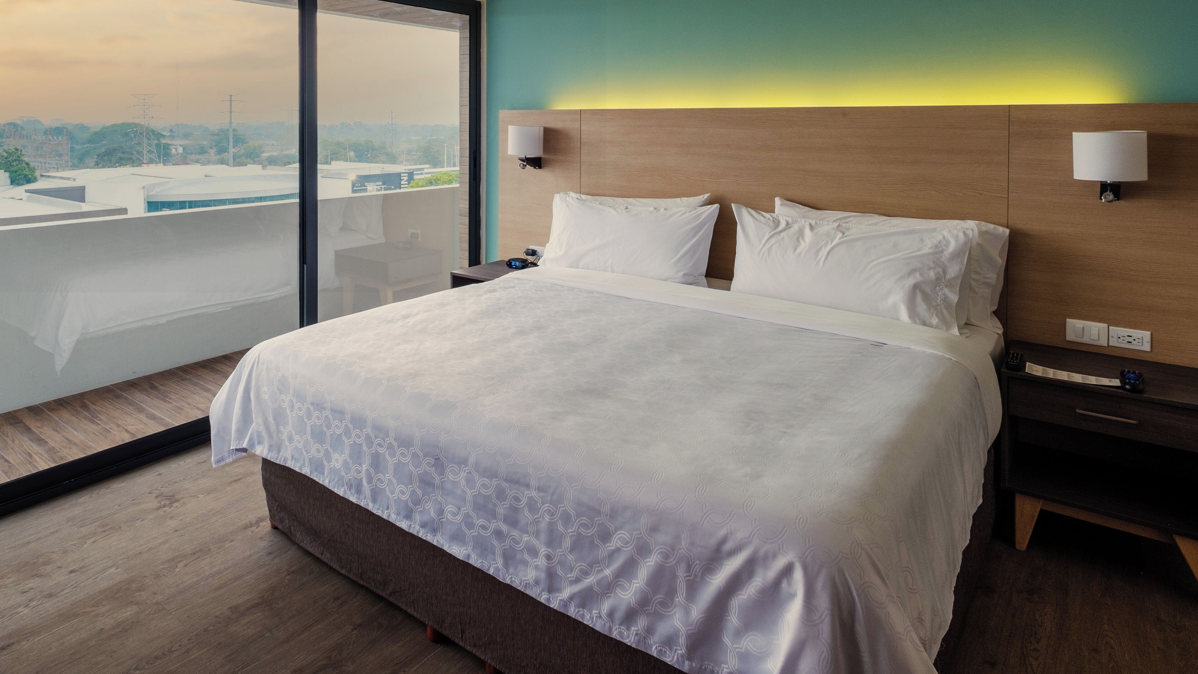 Habitación de hotel con una cama grande, lámparas de noche, mesita de noche y vista a la ciudad a través de un ventanal amplio.