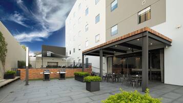 A modern outdoor patio with a covered seating area, outdoor kitchen, and a view of a multi-story building.