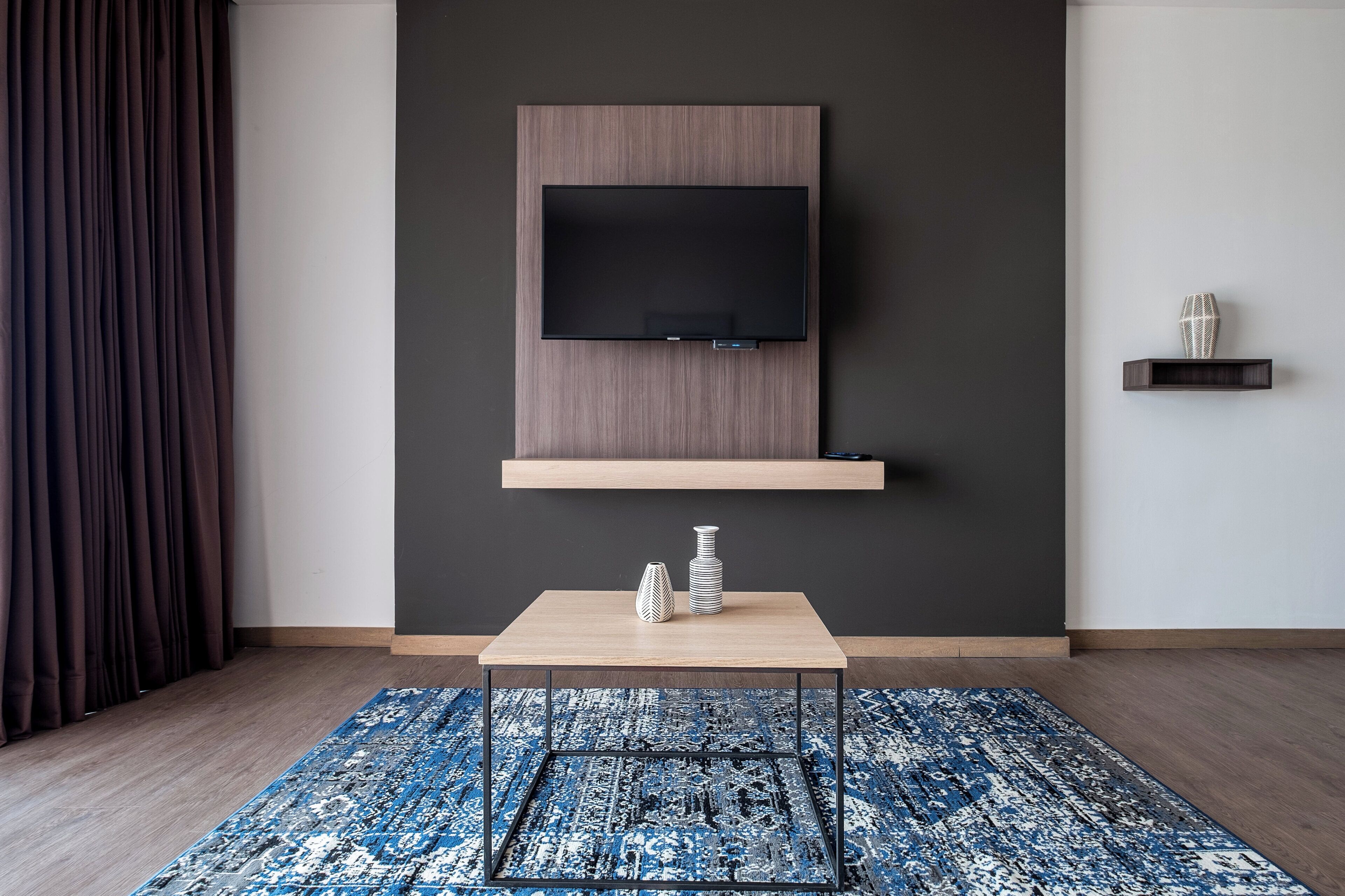 A modern living room with a mounted TV, a small wooden shelf, a table with decorative vases, and a patterned rug.