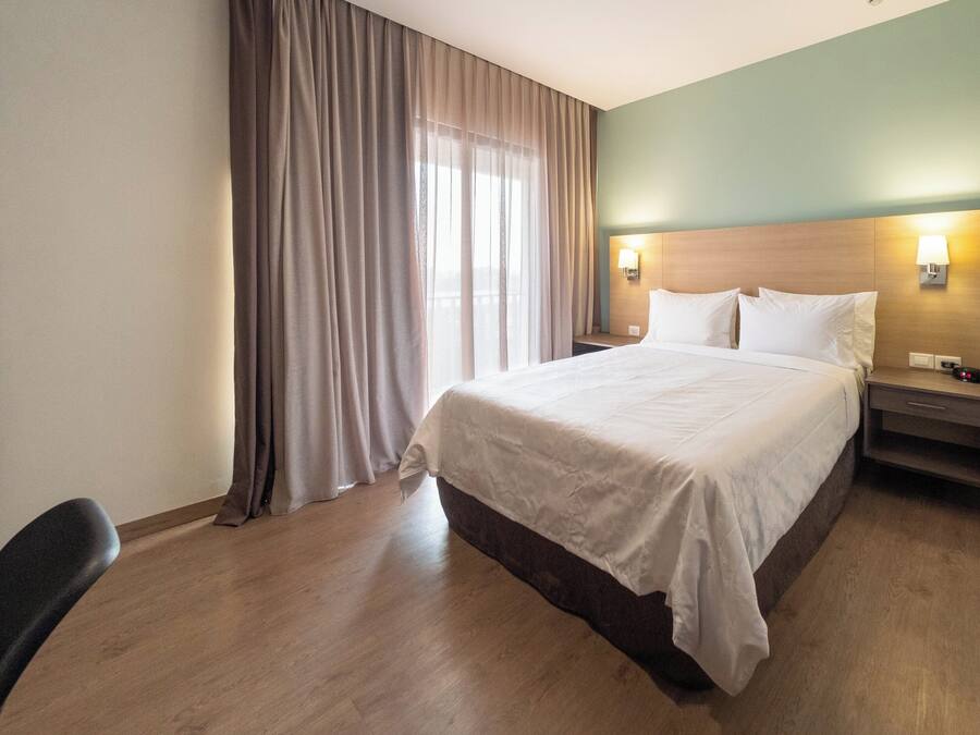 Una camera d'albergo con un letto, una scrivania, una sedia, un comodino e una finestra con tende.
