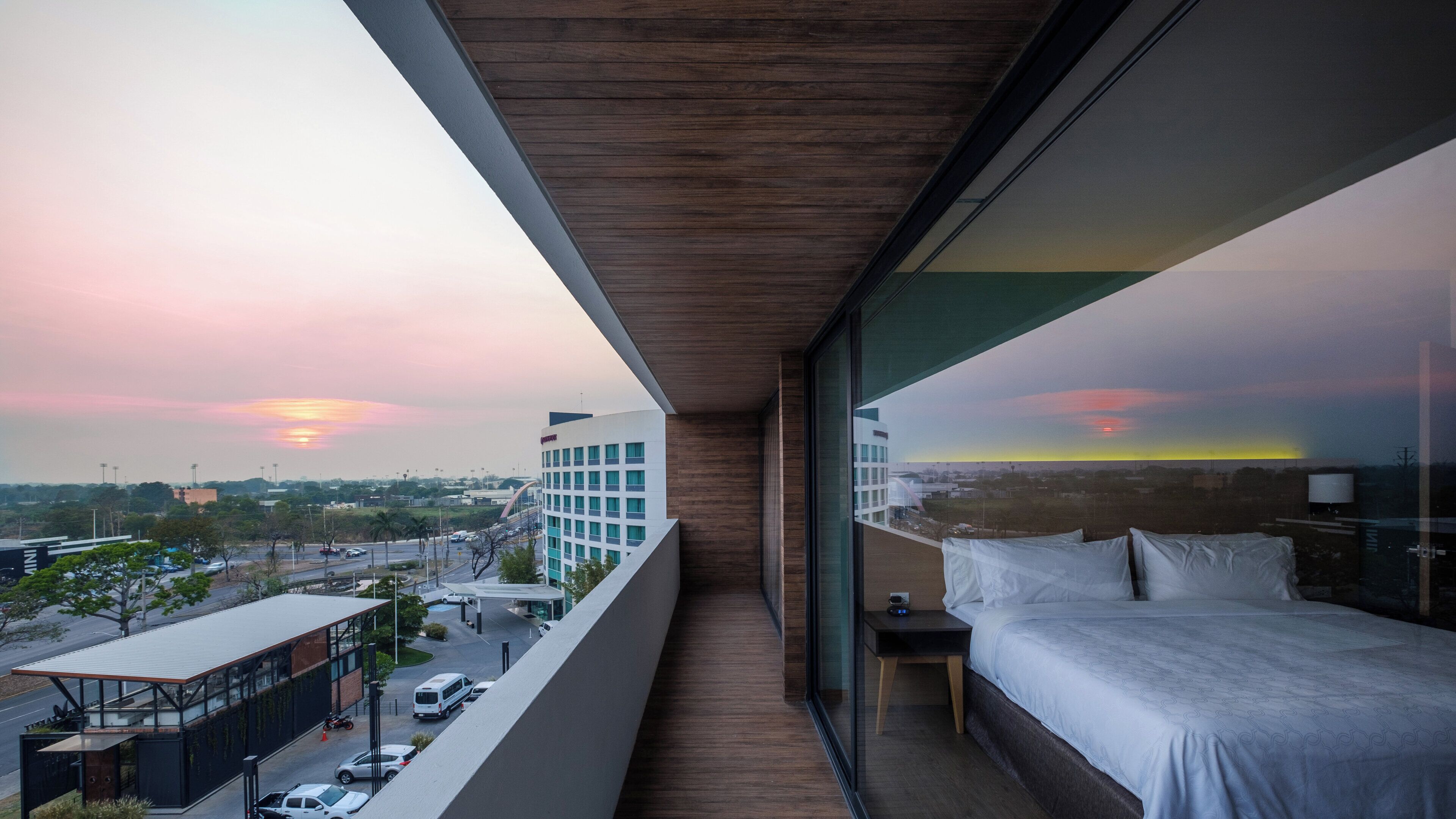 Un balcón con cama, mesita de noche y vista a la ciudad al atardecer.