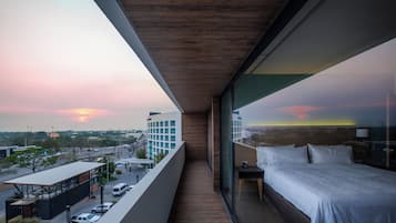 Un balcón con cama, mesita de noche y vista a la ciudad al atardecer.
