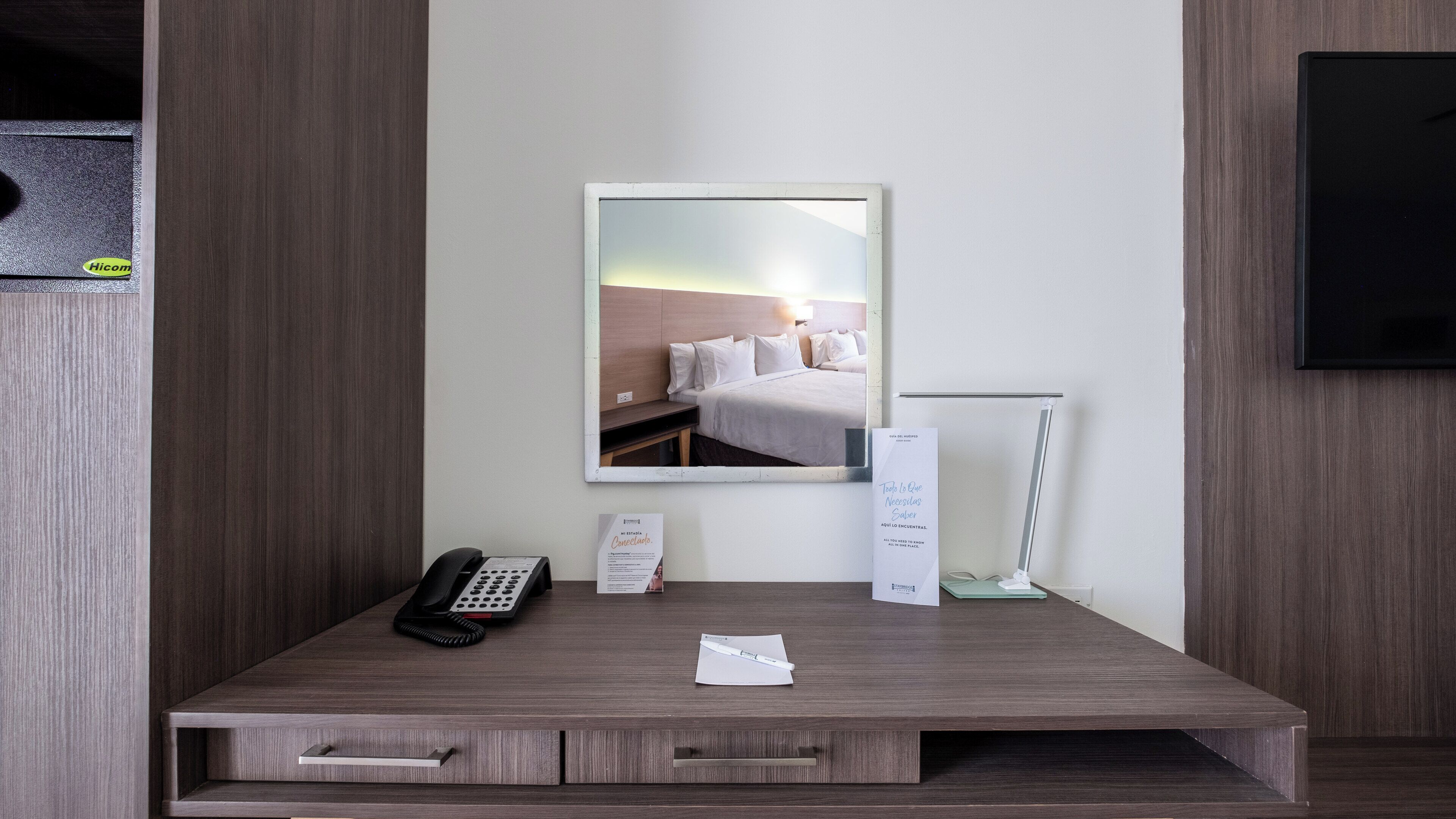 A hotel room with a wooden desk, a telephone, a mirror, a bed, and a wall-mounted TV.
