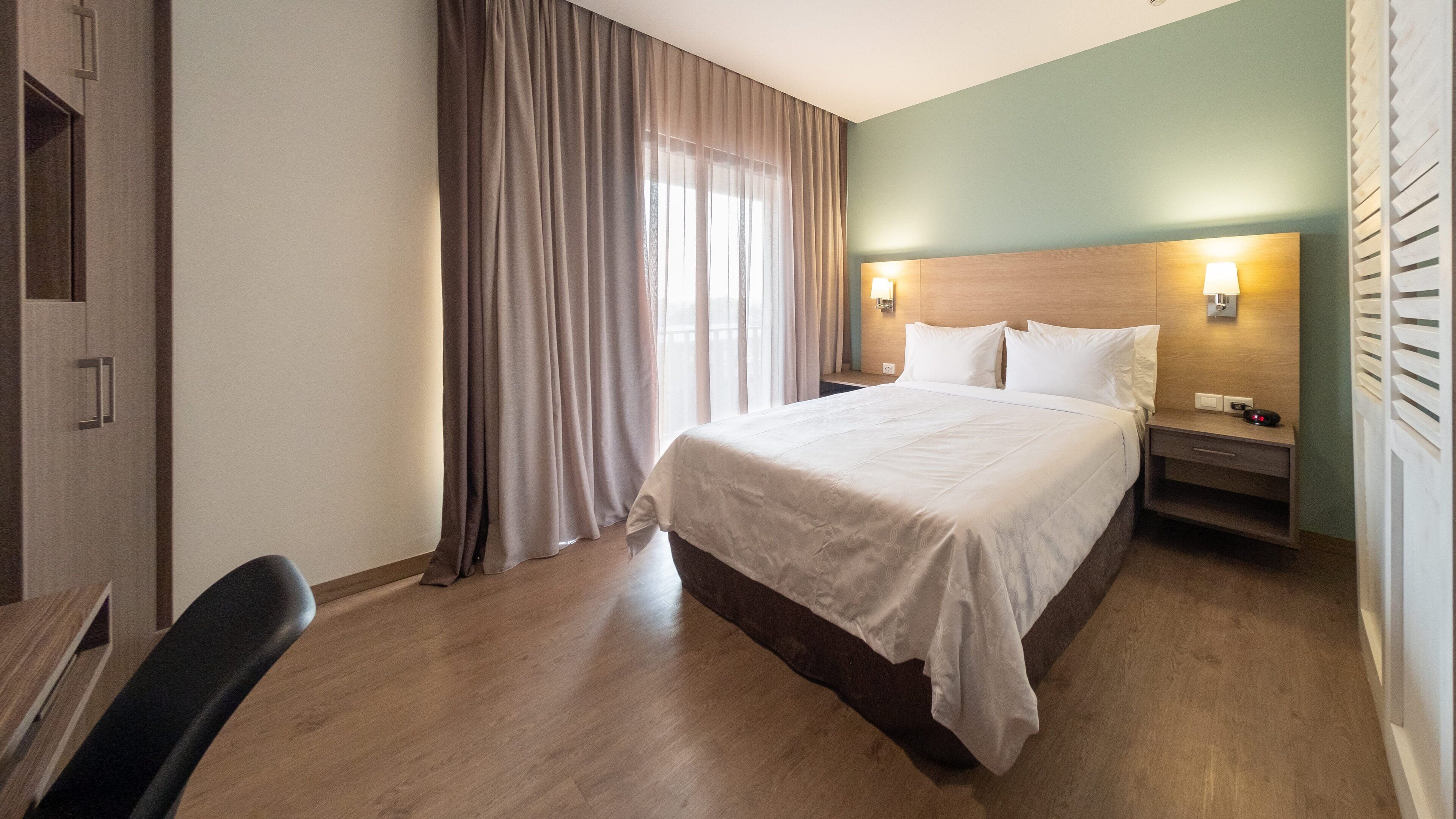Une chambre d’hôtel comprenant un lit, un bureau, une chaise, une table de chevet et une fenêtre avec des rideaux.