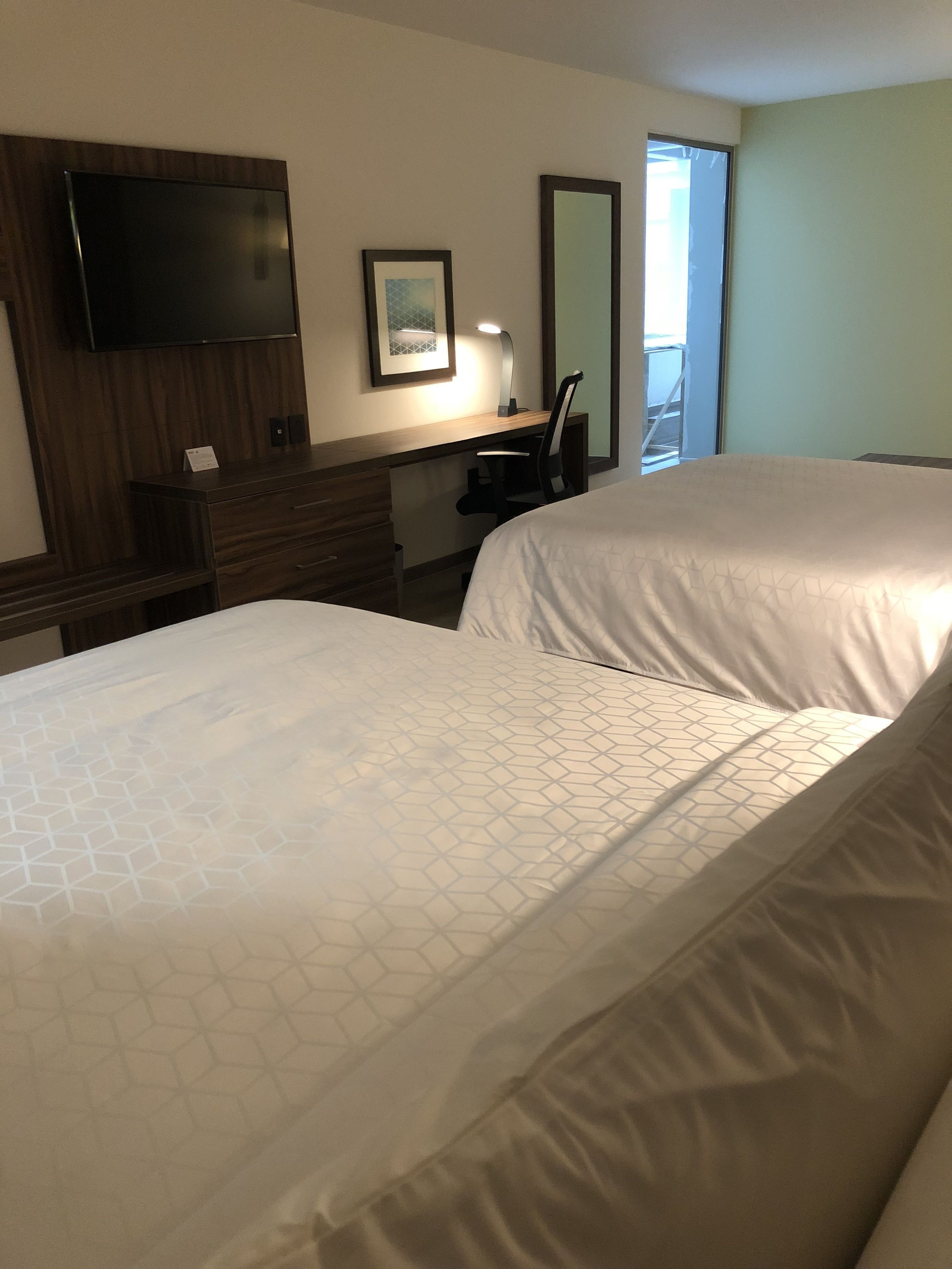 Habitación de hotel con dos camas, un escritorio con silla, televisor de pantalla plana montado en la pared, un cuadro enmarcado y una ventana con vistas.