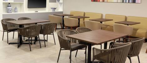 Interior de una cafetería moderna con varias mesas y sillas, un televisor de pantalla plana en la pared y jarrones decorativos en estantes.