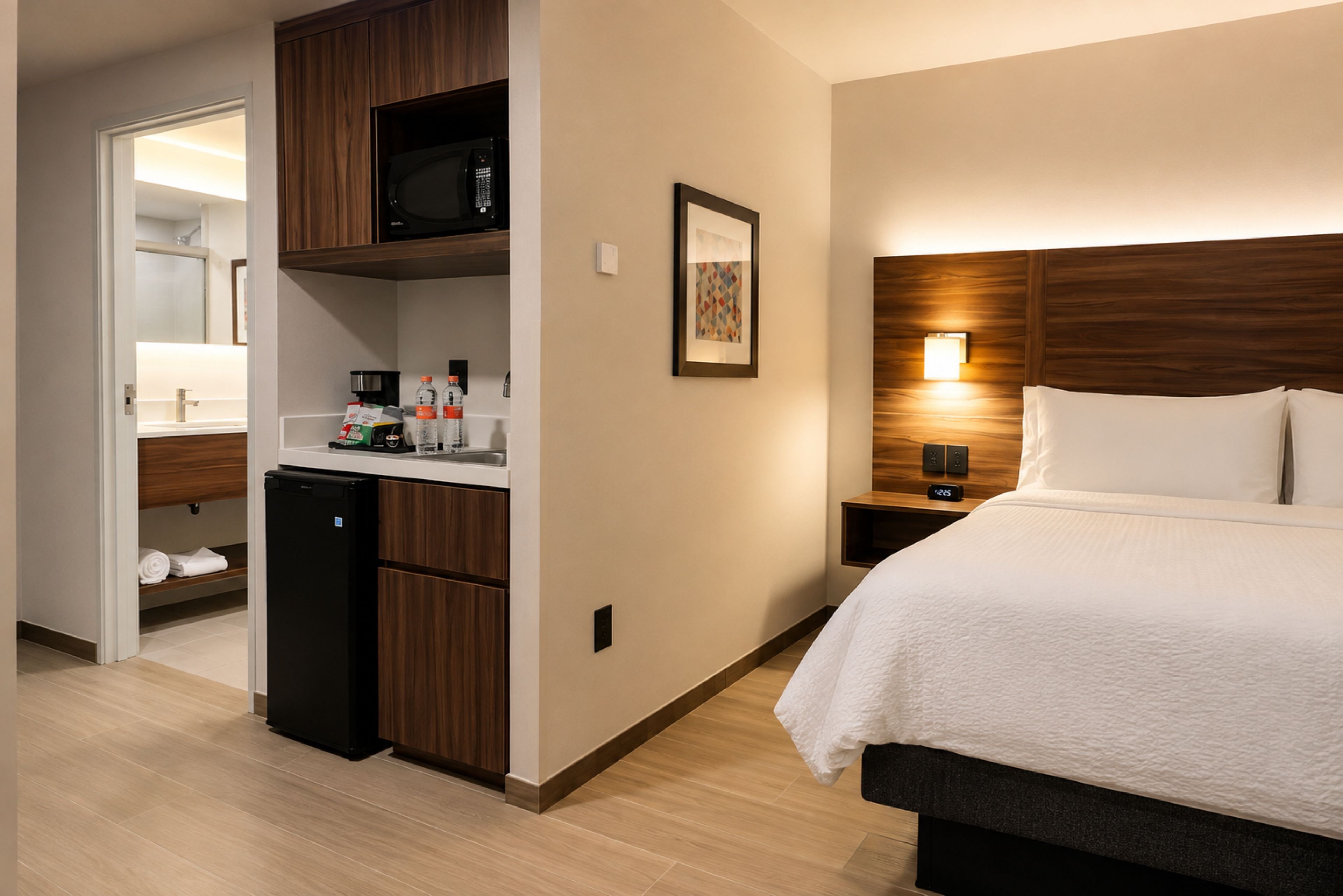 A hotel room with a bed, a small kitchenette, a microwave, a refrigerator, a bedside table with a lamp, and a framed picture on the wall.