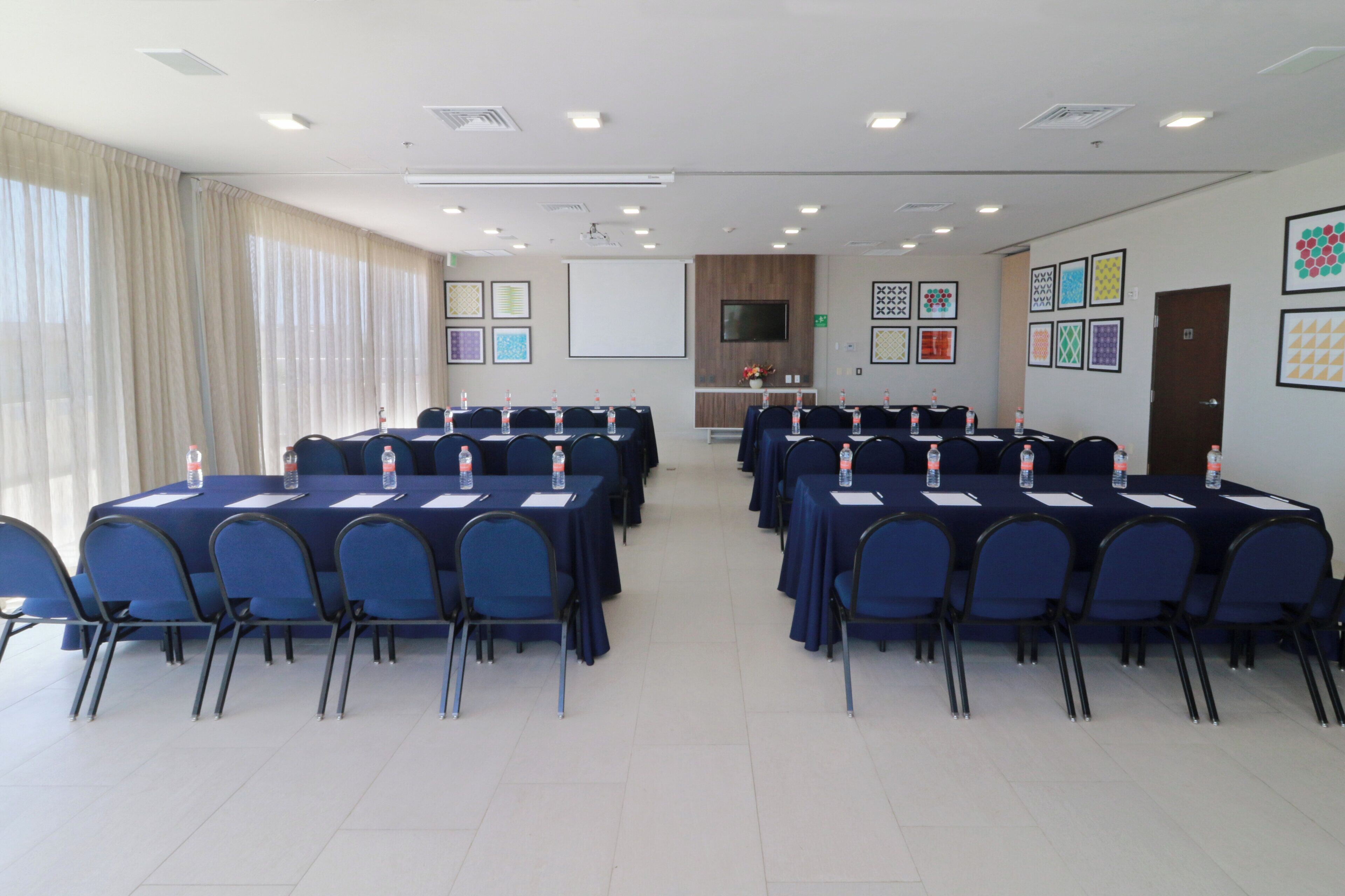 Un salón de conferencias con varias mesas y sillas, una pantalla de proyector y un televisor en la pared.