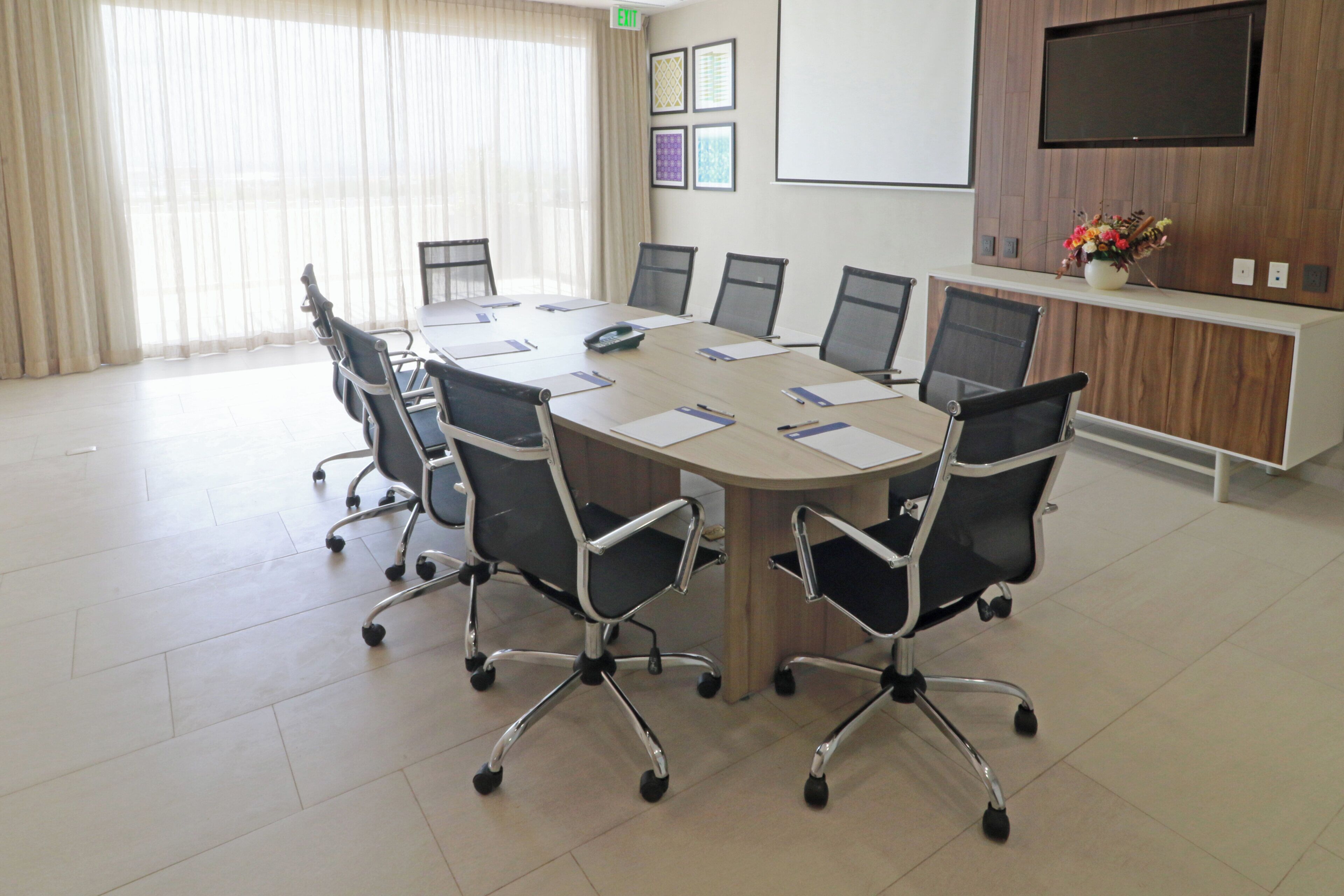 A conference room with a long table, chairs, a projector screen, and a wooden cabinet with flowers.
