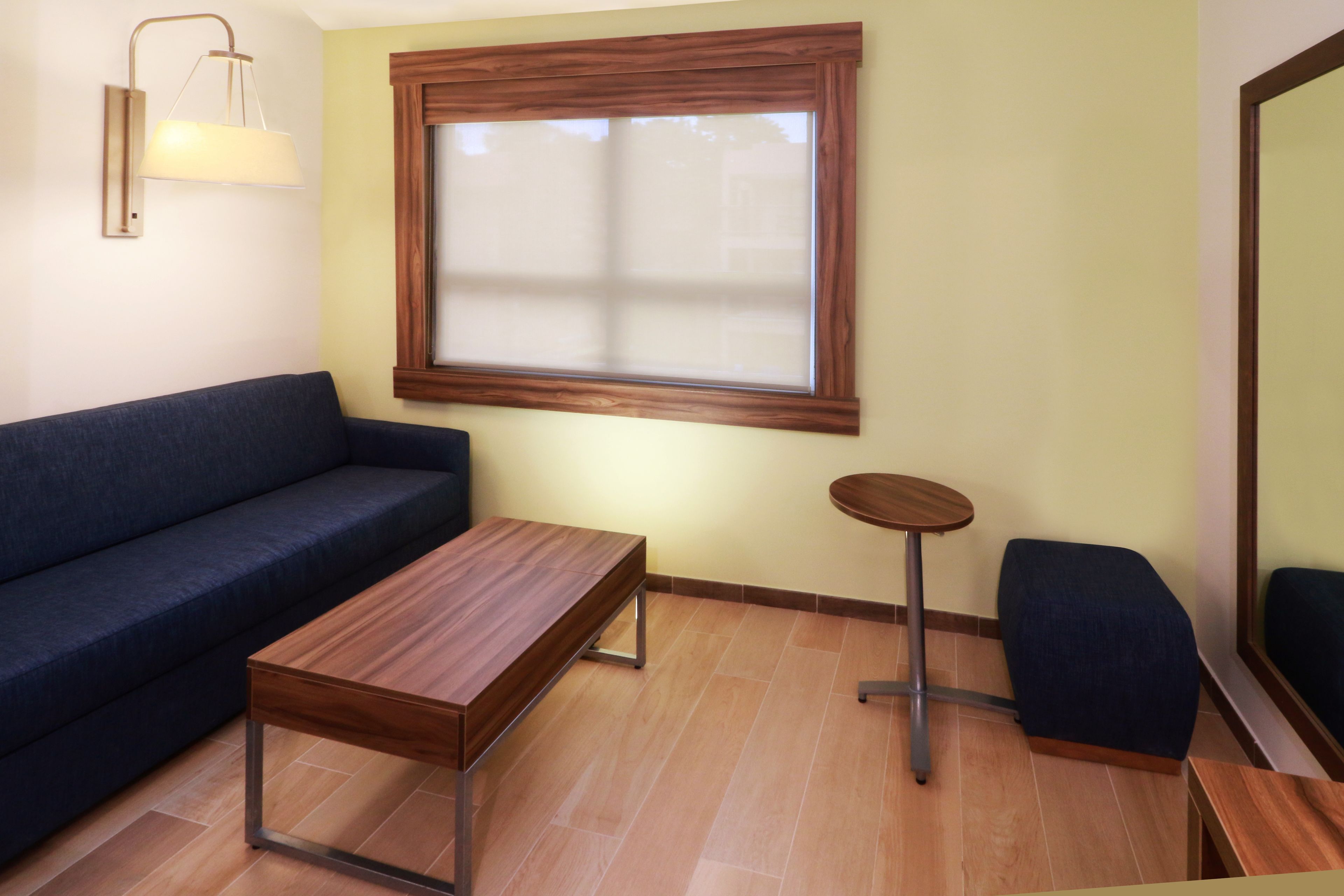 Um quarto com um sofá azul escuro, uma mesa de centro de madeira, uma mesa redonda pequena e uma janela grande com persianas.