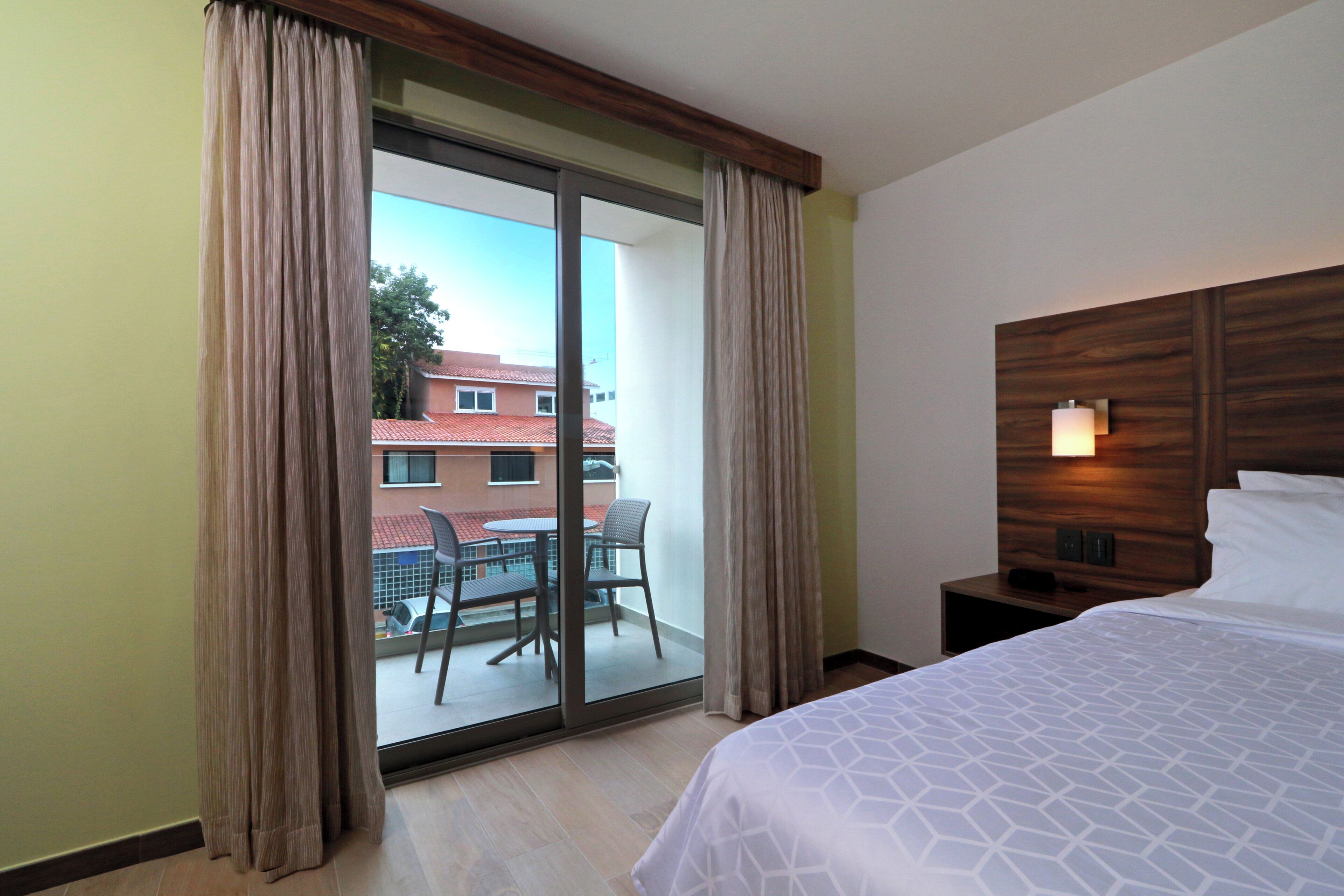 A hotel room with a bed, a nightstand, a lamp, a sliding glass door to an outdoor area with a table and chairs, and a view of buildings outside.