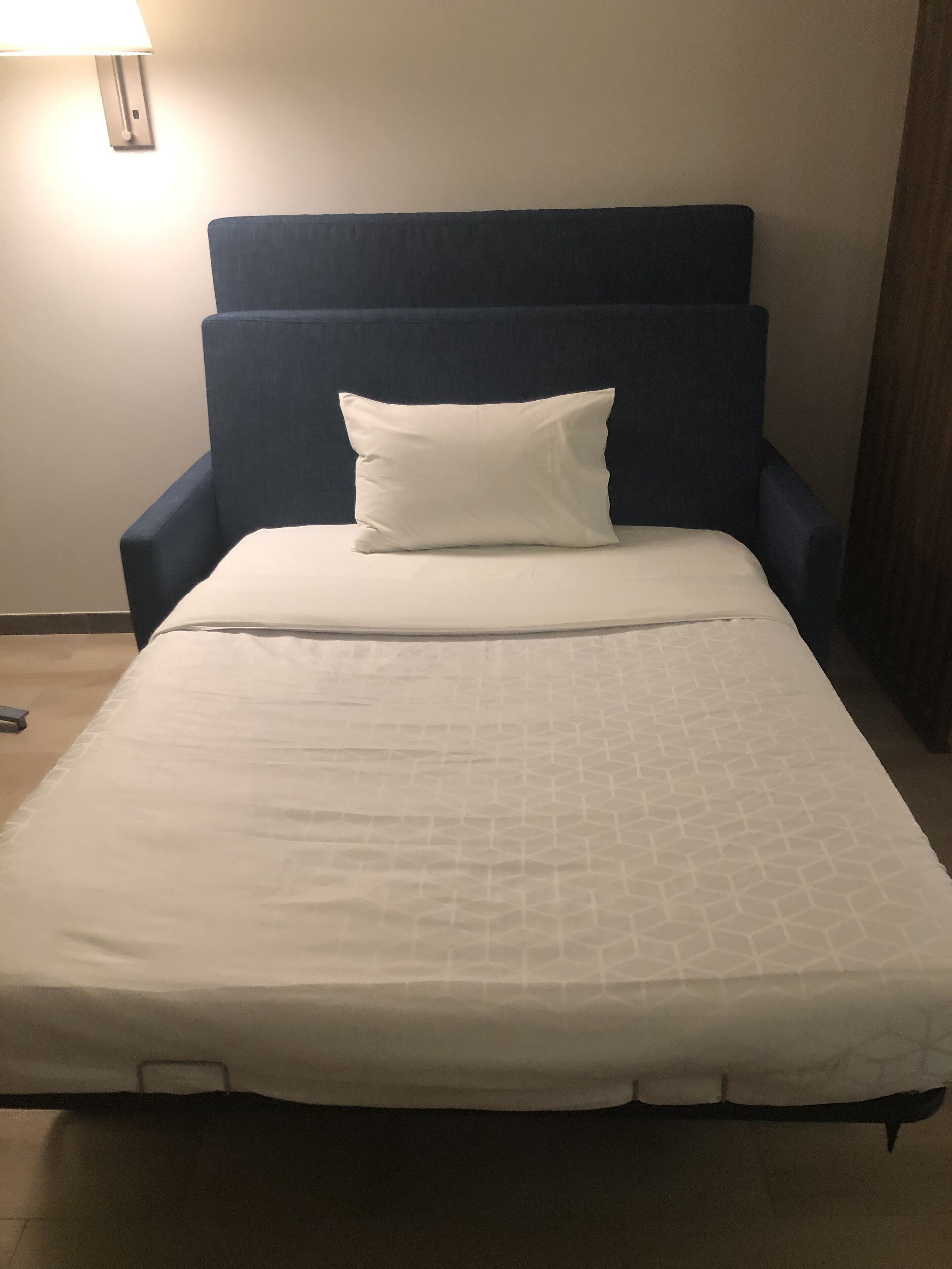 Una cama individual con colchón y almohada blancos, cabecero azul oscuro y una lámpara fijada a la pared.