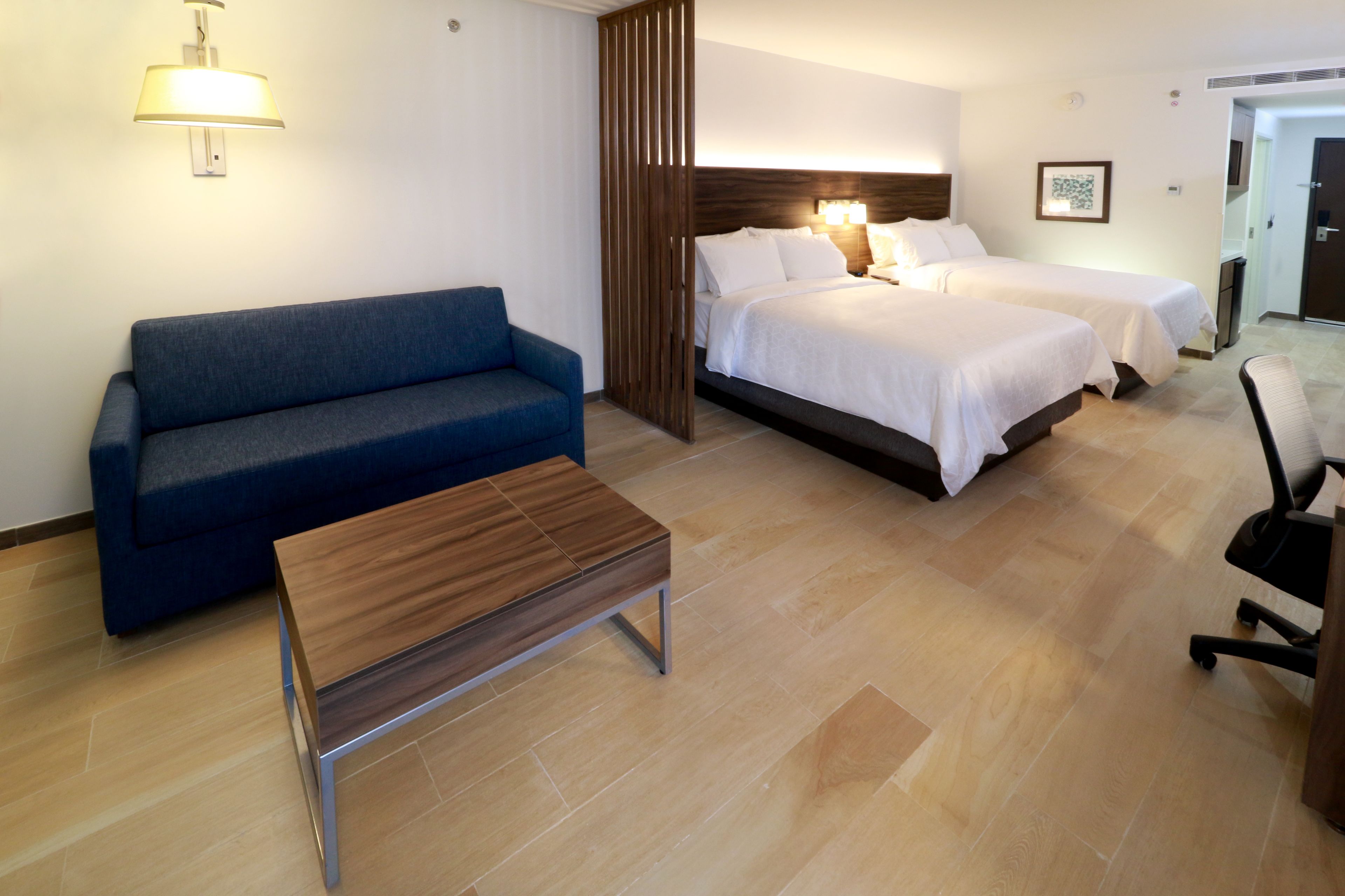 A hotel room with a blue sofa, a wooden coffee table, two beds, and a desk with a chair.