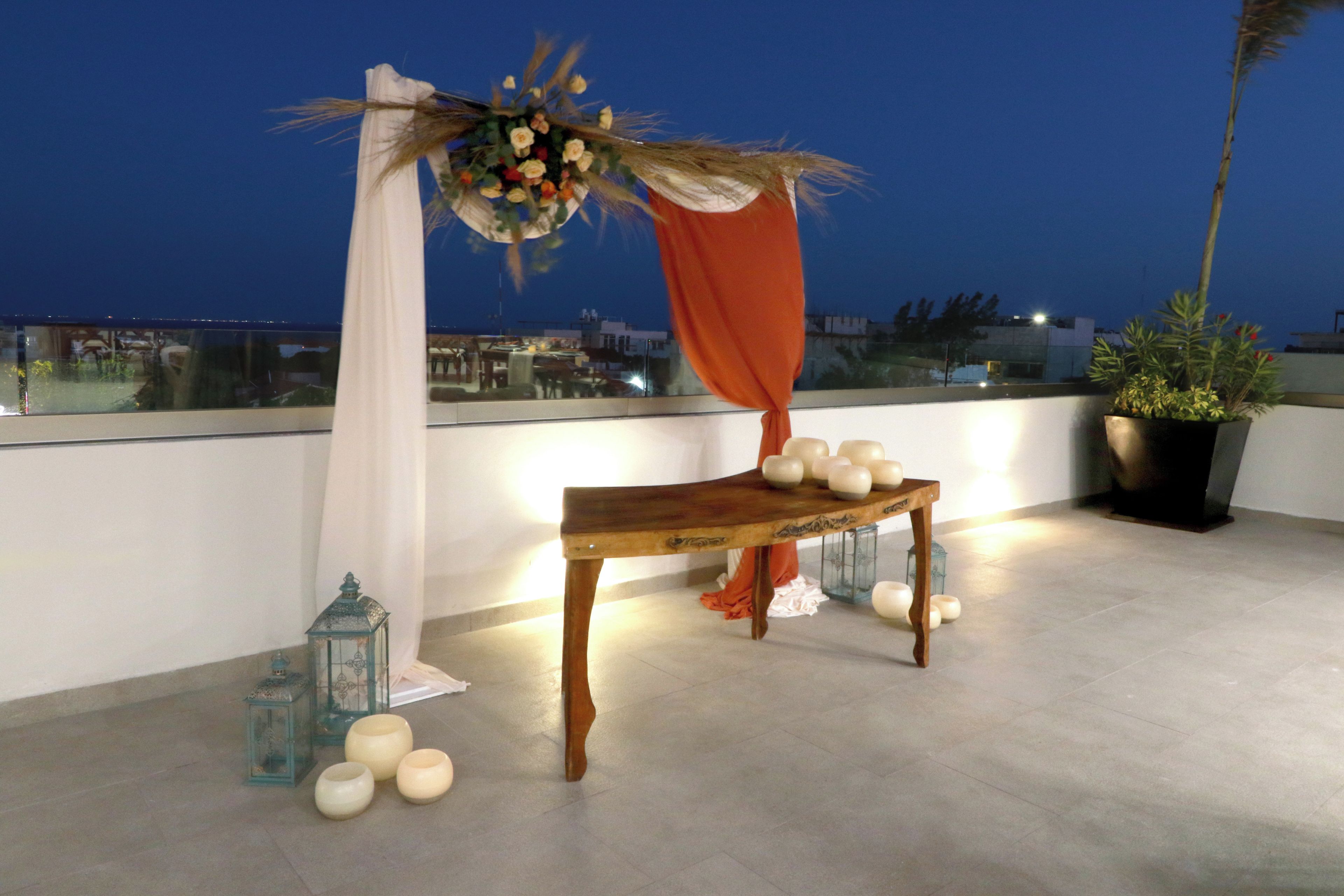 A rooftop terrace with a wooden table, white candles, and a decorative arrangement with fabric and flowers.