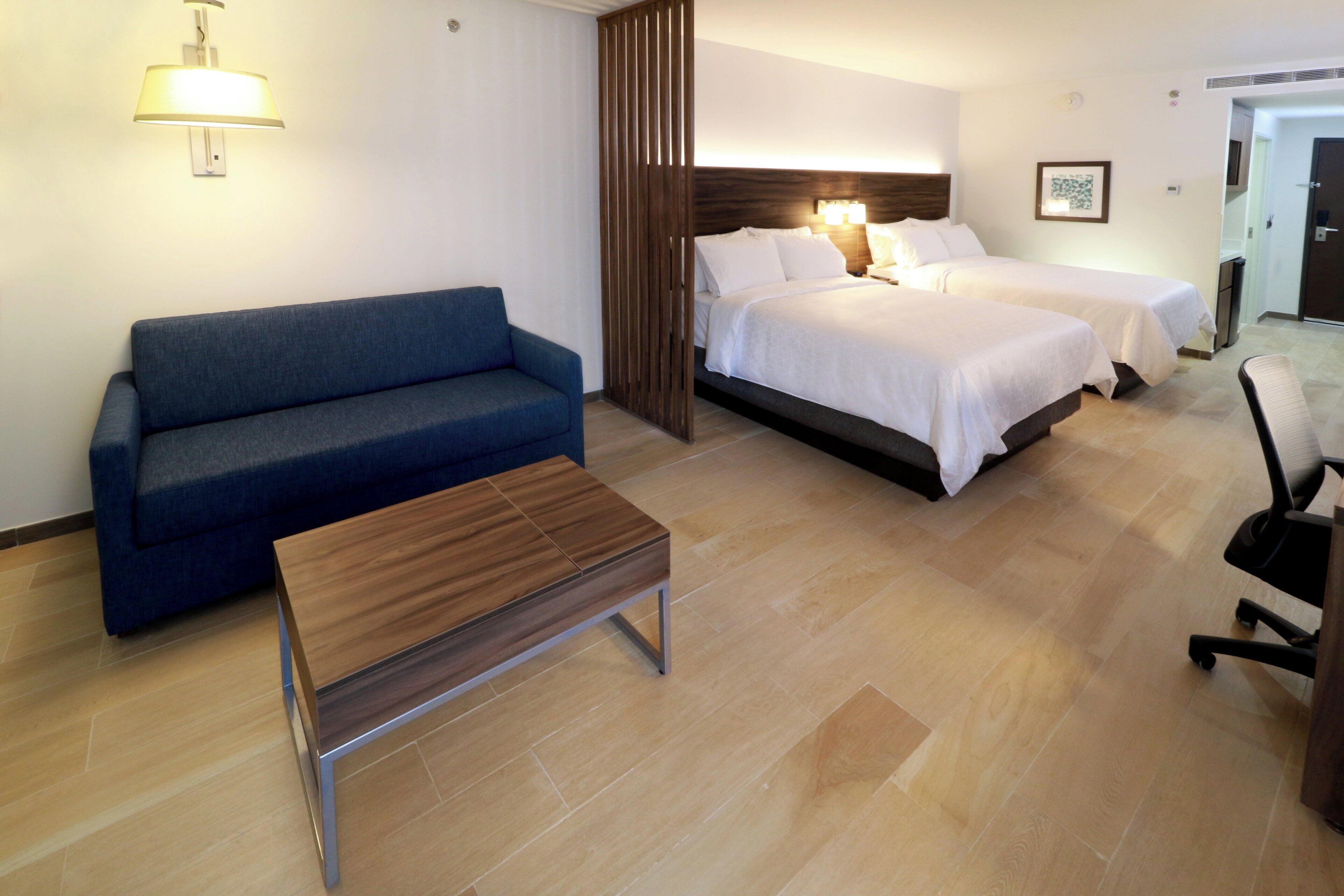 A hotel room with a sofa, two beds, a small table, and a lamp.