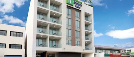 A Holiday Inn Express & Suites building with a modern design, featuring a flat roof, multiple balconies with glass railings, and a clear sky in the background.