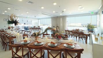 Un comedor amplio con varias mesas dispuestas para una comida, ventanas grandes con vistas y un diseño moderno de techo.