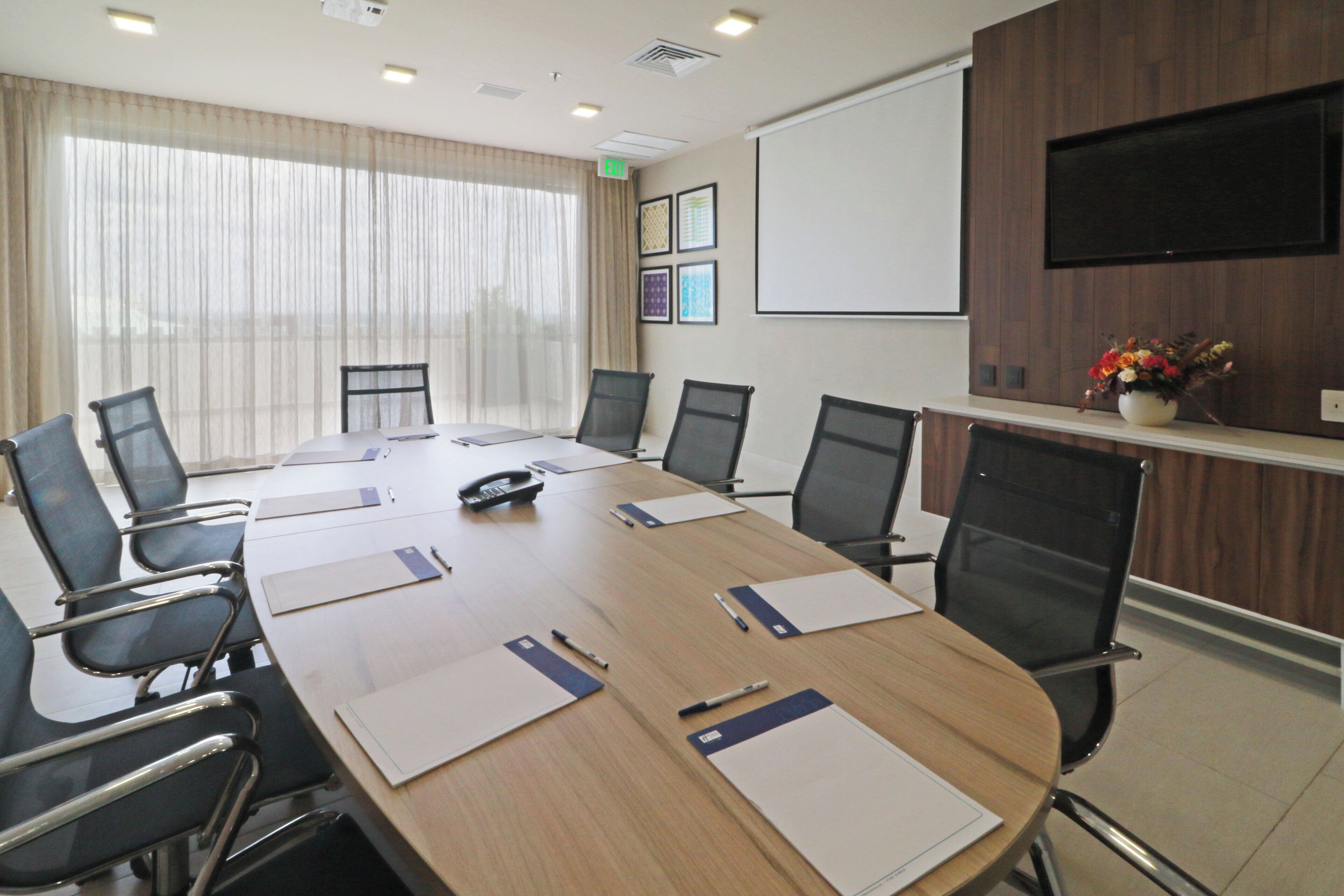 Un salón de conferencias con una mesa larga, sillas, una pantalla de proyector y un televisor en la pared.