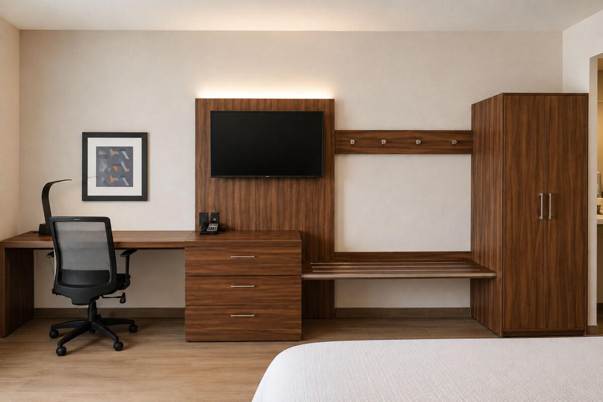 Ein Hotelzimmer mit einem Holztisch, einem schwarzen Bürostuhl, einem flachbildfernseher an der Wand, einem Bett mit weißer Bettwäsche und einem zweitürigen Kleiderschrank.