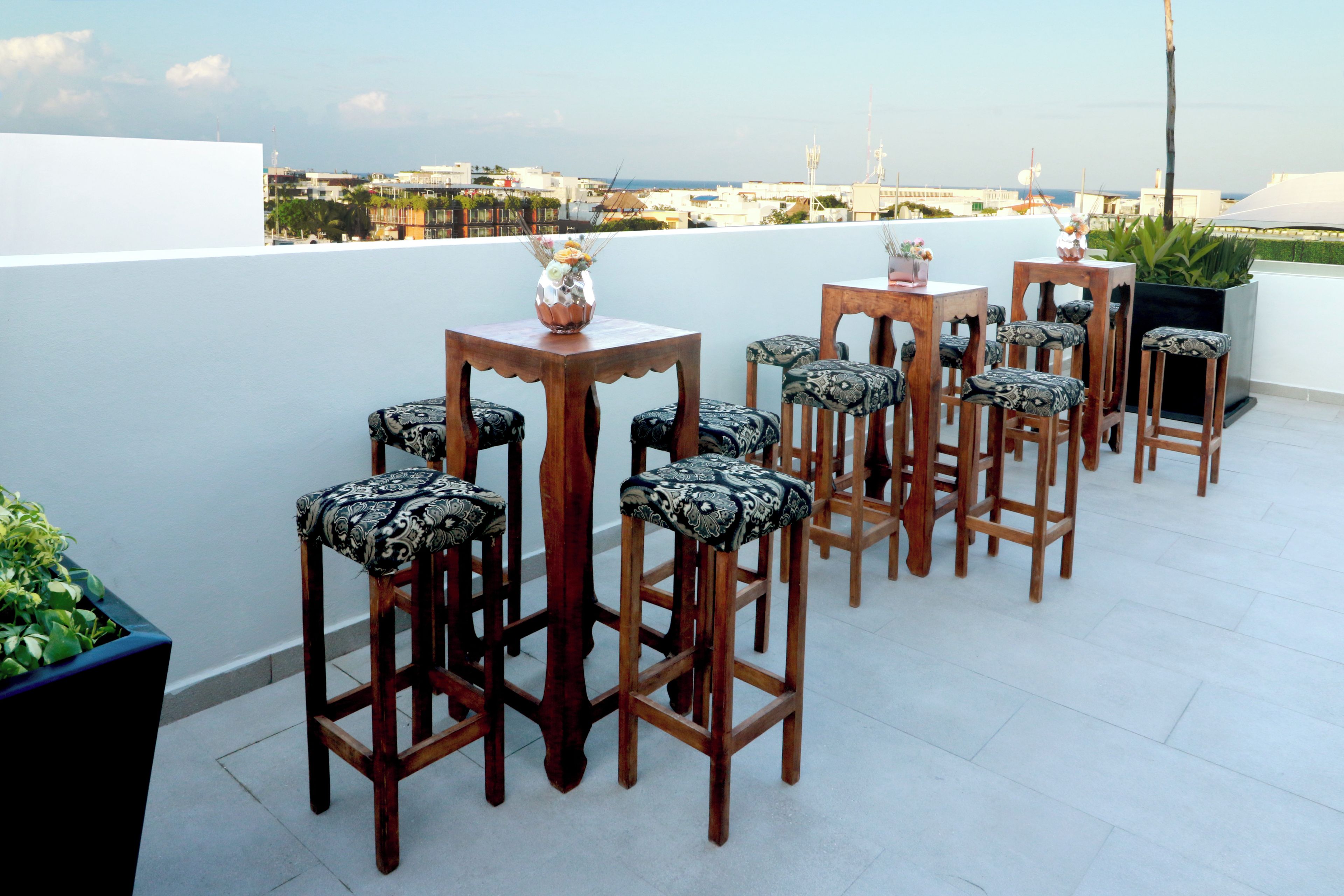 A rooftop terrace with wooden bar stools and tables, a potted plant, and a view of the city skyline.