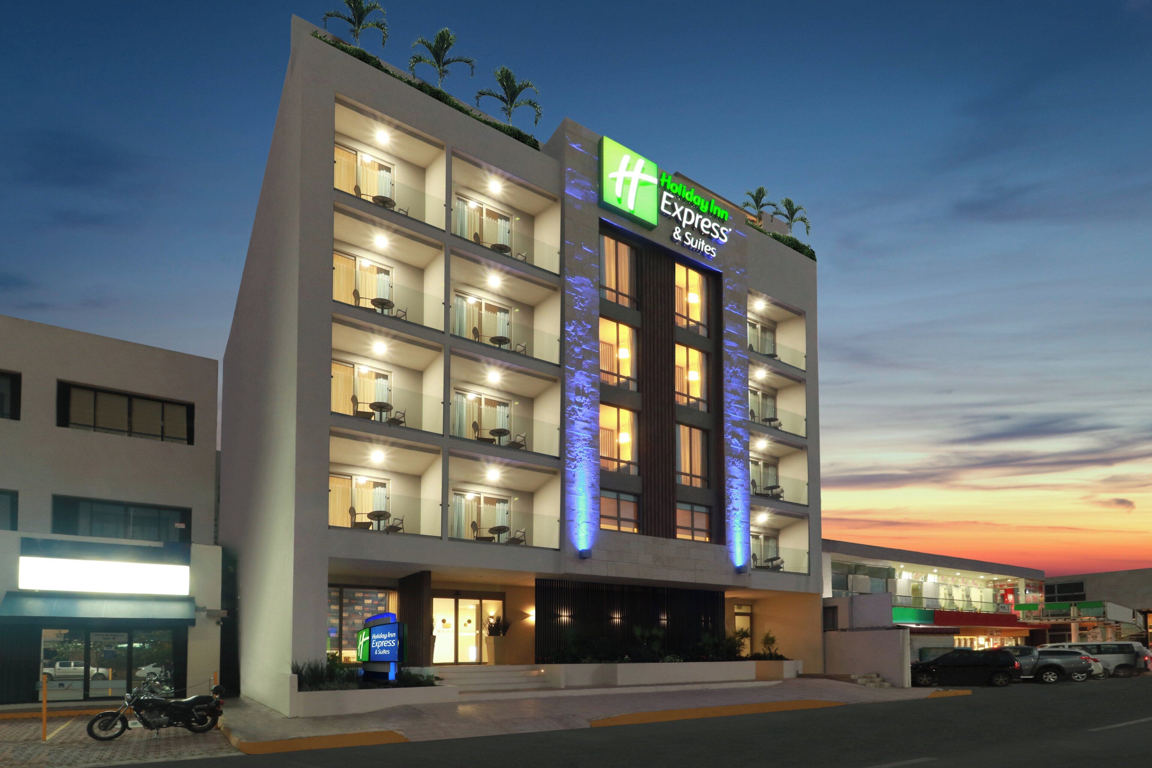 A multi-story hotel with a prominent Holiday Inn Express sign, balconies with seating, and a parking area with vehicles.