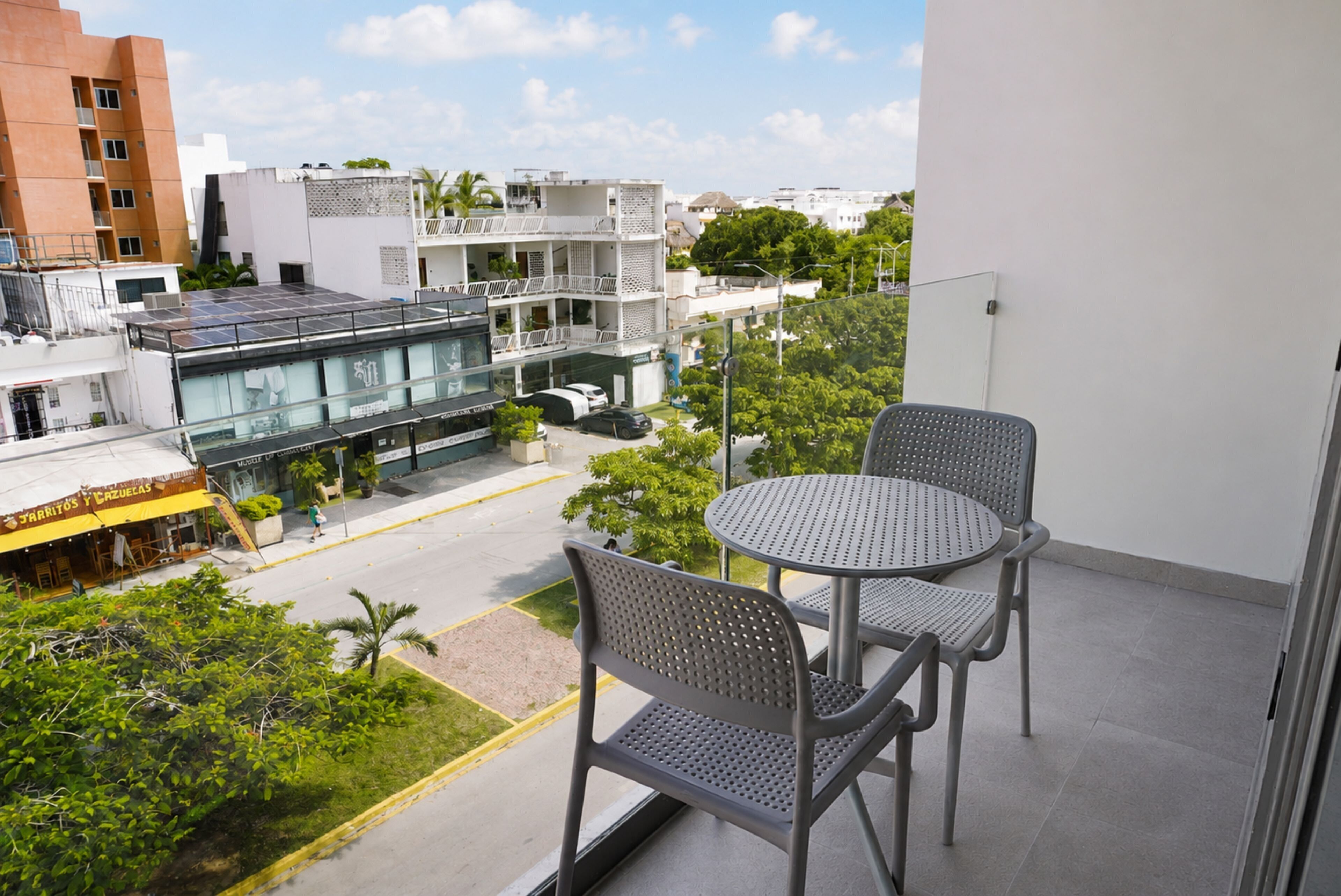 A balcony with two metal chairs and a small table, overlooking a street with buildings and a restaurant.